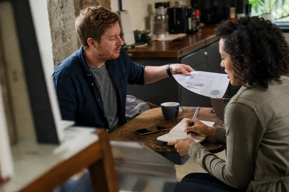Two colleagues reviewing a customer account brief side by side at a cafe table inside an office